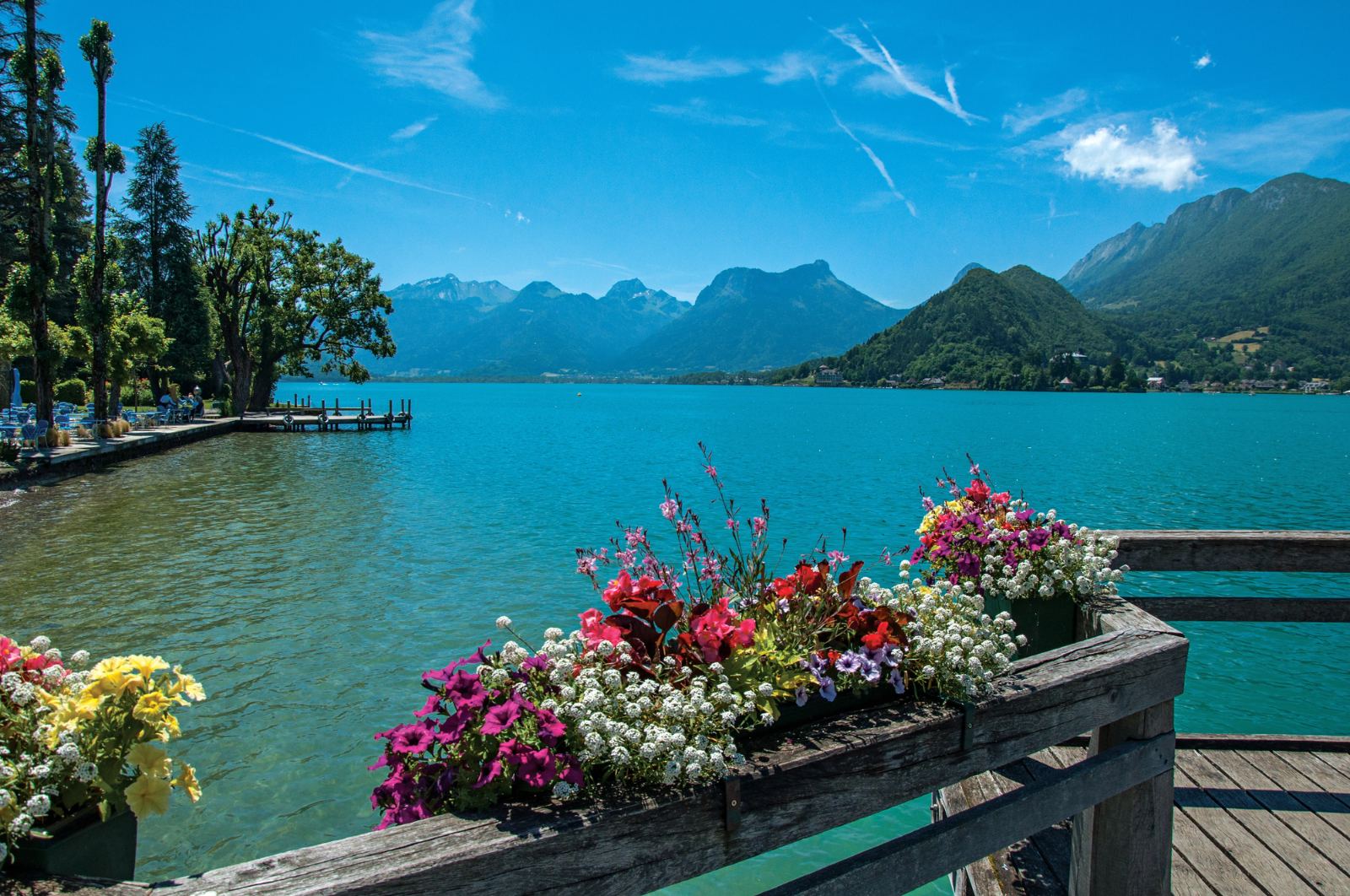 Flower boxes on a wooden pontoon over the turquoise water of Lake Annecy with mountains behind