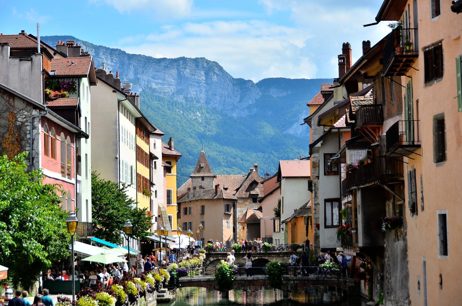 Canal in old Annecy lined with pastel facades, flowers and mountains behind